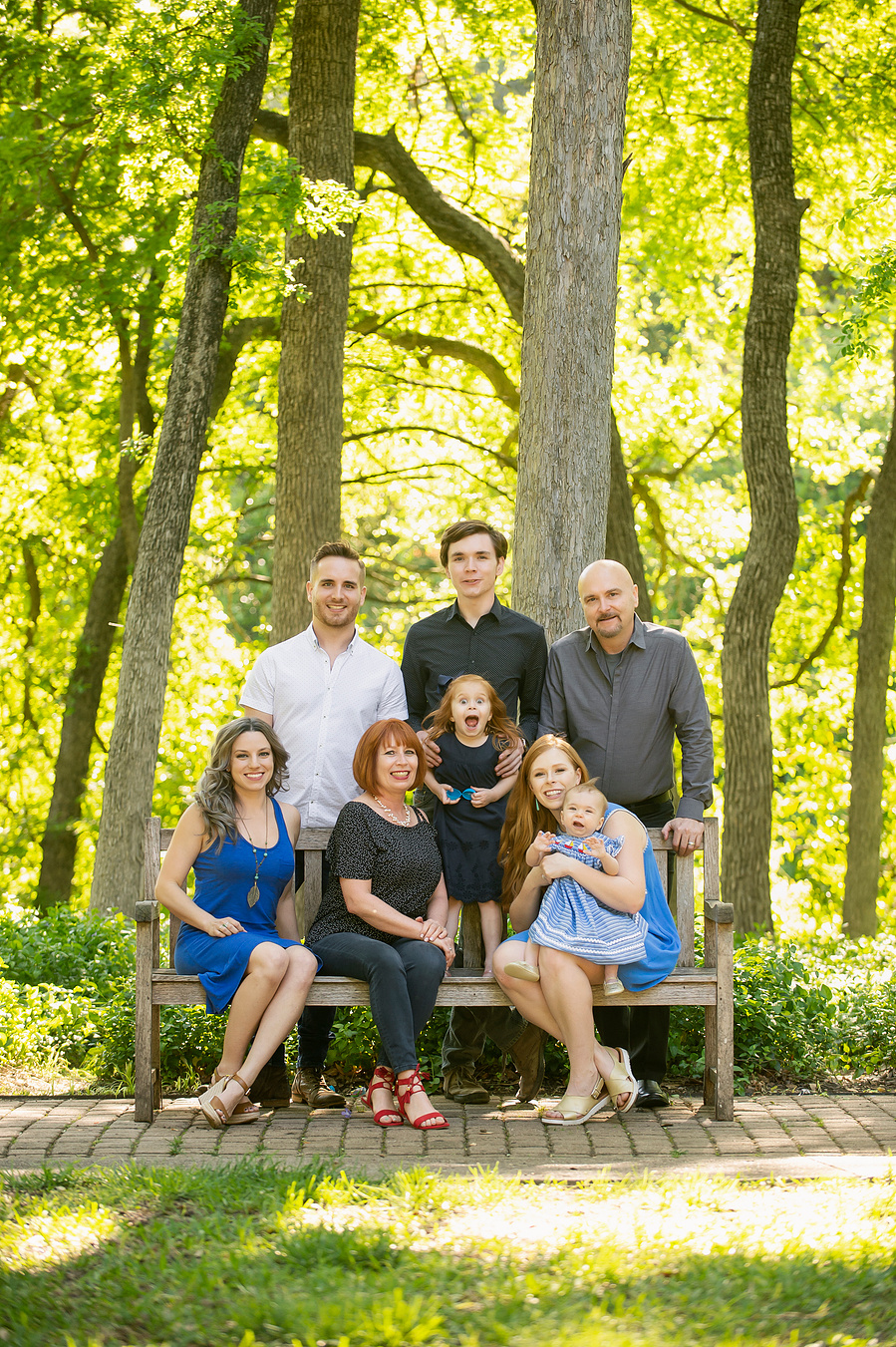 A multi-generational extended family gathered on a bench in a shaded Dallas park