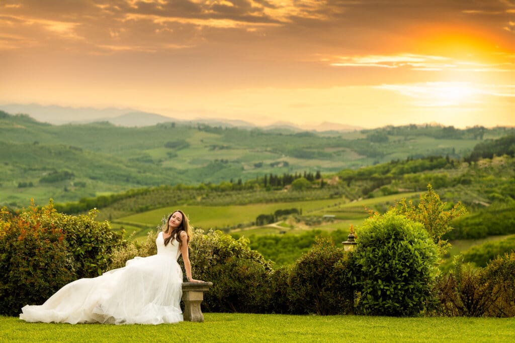 Bride sitting on a stone garden bench with a vibrant, glowing sunset over the hills of Tuscany in the background.
