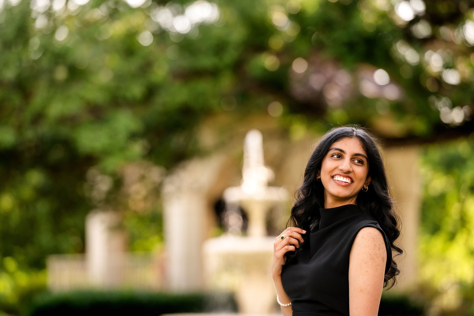 A smiling senior in a black dress in front of a garden fountain in Dallas