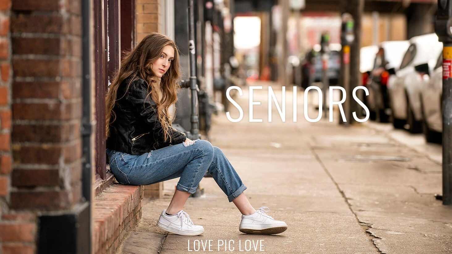 Magazine-quality urban senior portraits in Dallas, TX. A high school senior girl wearing a black leather jacket sits on a city storefront ledge for a luxury, unposed Class of 2026 photography session.