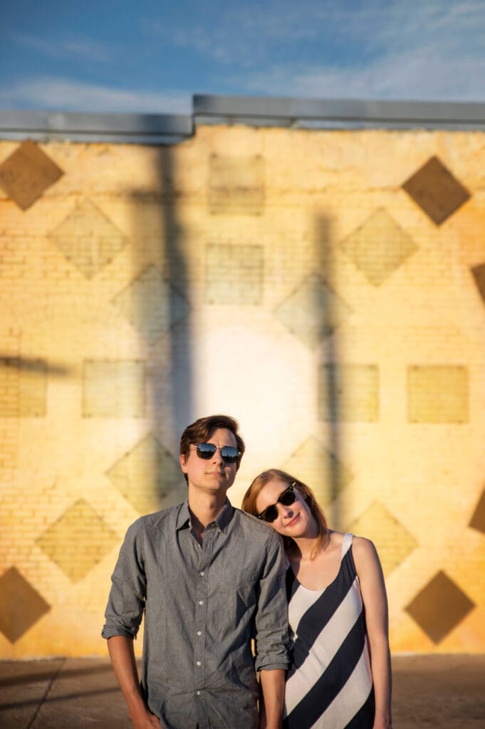 Couple posing in front of a geometric yellow brick mural wall in Dallas during golden hour, both wearing sunglasses