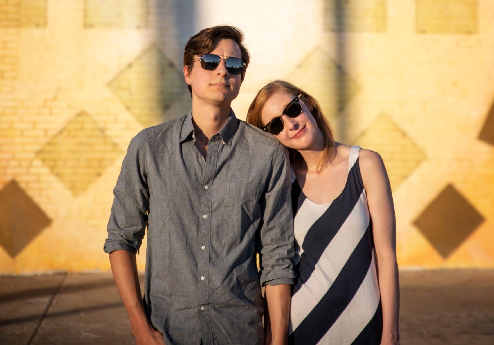 Couple posing in front of a geometric yellow brick mural wall in Dallas during golden hour, both wearing sunglasses