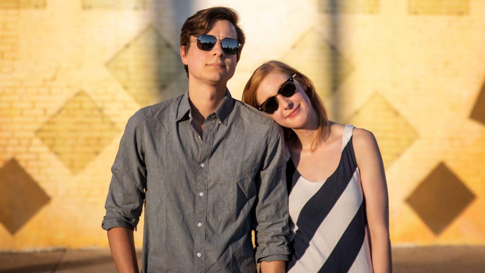 Couple posing in front of a geometric yellow brick mural wall in Dallas during golden hour, both wearing sunglasses