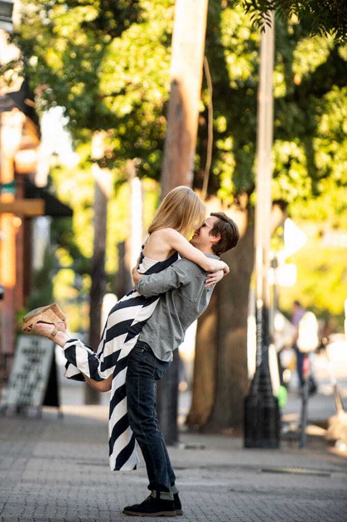Man dipping his partner during an engagement session on a Deep Ellum street surrounded by trees and urban architecture