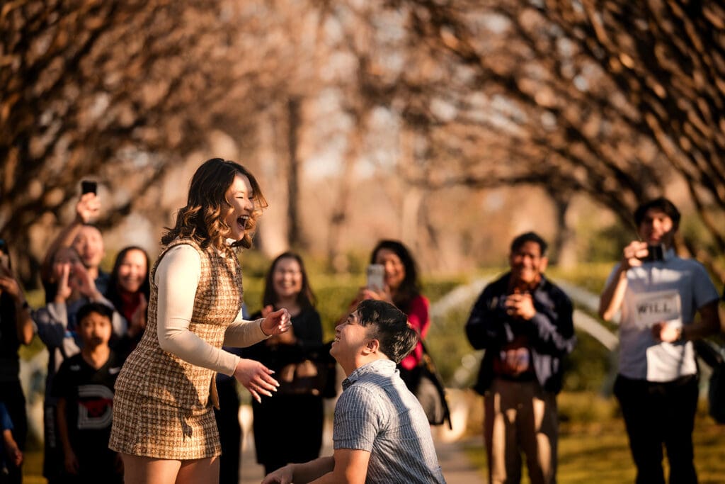 Candid reaction of a woman being proposed to at the Dallas Arboretum while an excited crowd watches in the background.