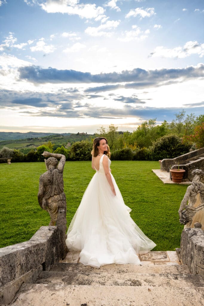Bride walking down ancient stone steps past garden statues at a villa in Tuscany, looking back over her shoulder.
