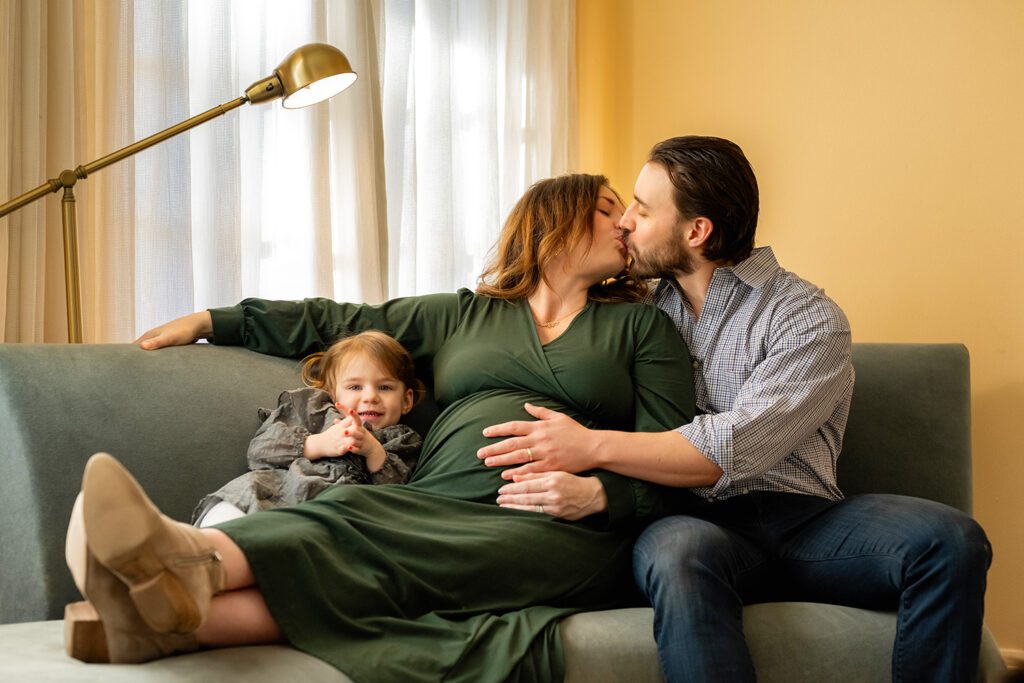 In-home maternity and family photography session in Dallas featuring a pregnant mother, father kissing, and a toddler sitting on a living room couch.