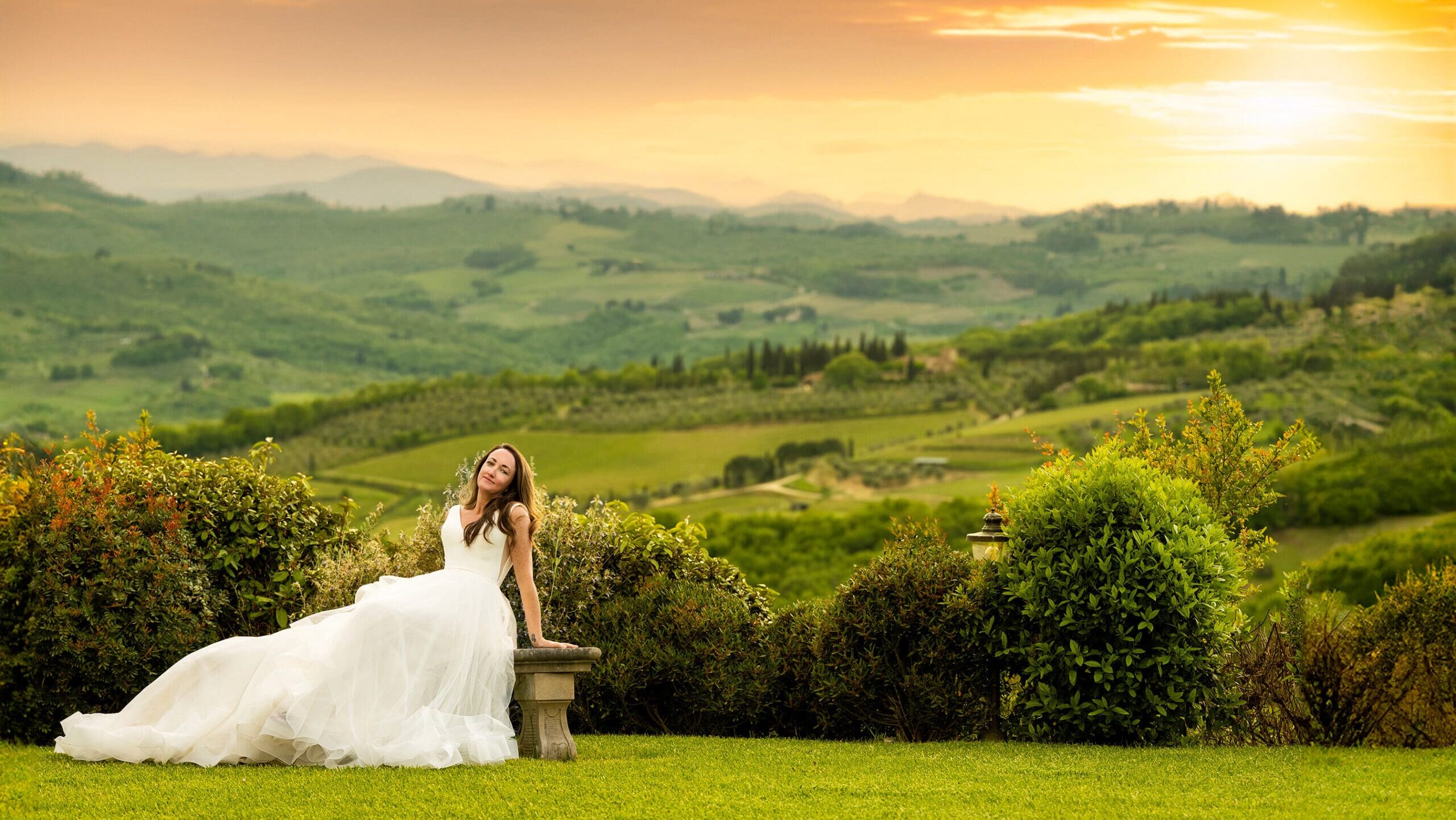 Bride in a flowing white wedding gown standing on a green lawn overlooking the rolling hills of Tuscany.