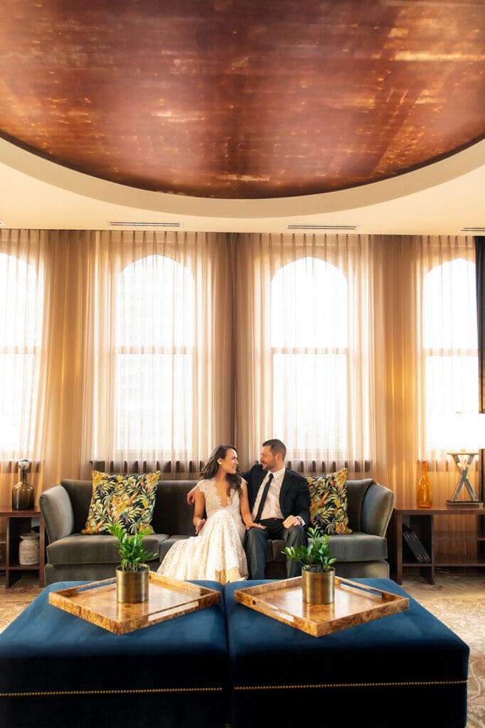 Bride and groom sitting on plush blue velvet couches under architectural ceilings in The Joule Dallas penthouse.